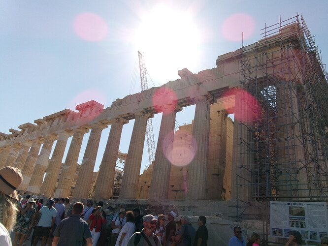 the many tall marble columns of the side of the Parthenon, with crowds of people surrounding the area along with construction equipment and scaffolding visible around the columns. There is a distinct lens flare. This photo is not labeled